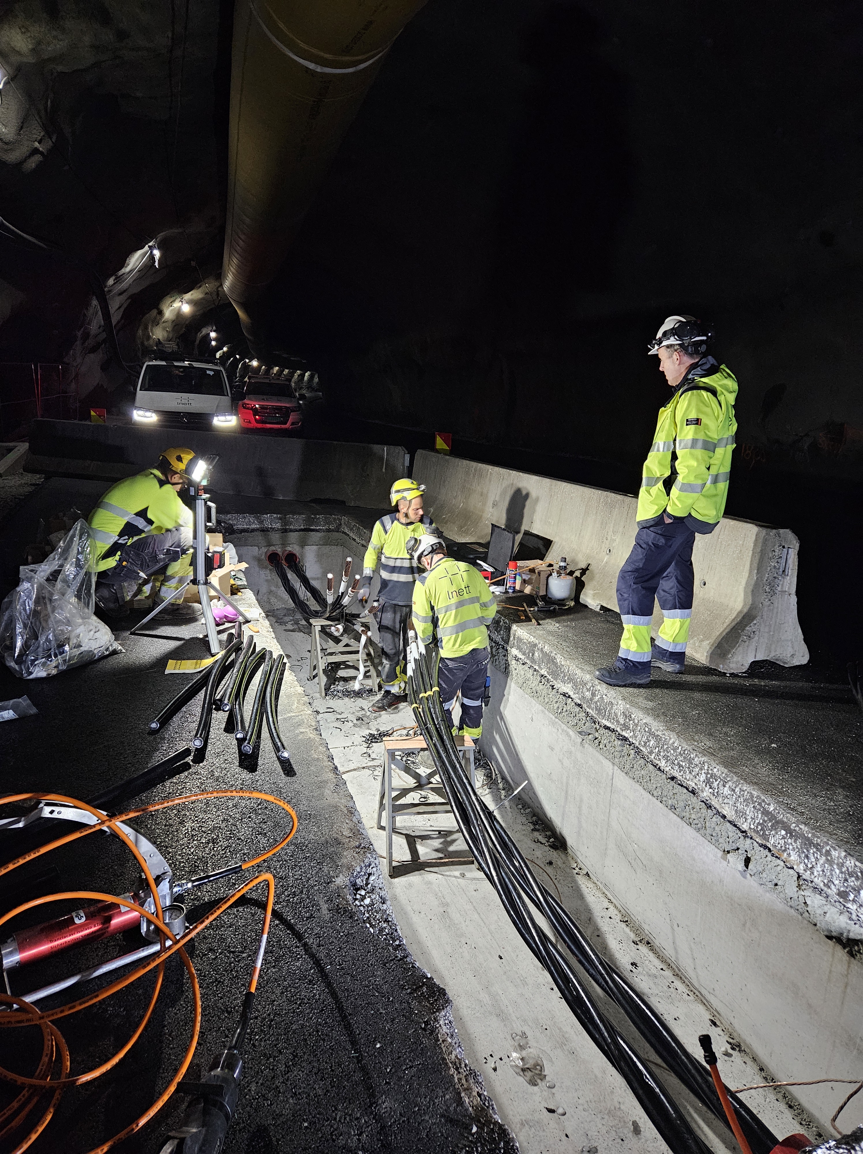 To montører står i betongstøpt grop og skjøter kabler inne i mørk tunnel, mens prosjektleder står ved siden av og hører hvordan arbeidet går. 
